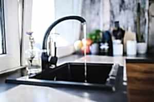 Close-up photo of a kitchen sink with a single-handled faucet stuck in the "on" position, water flowing from the spout. This image depicts a situation where you might need to follow a guide on how to remove a stuck tap handle.
