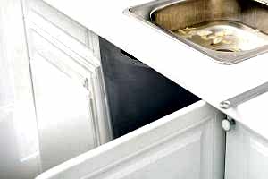 Kitchen sink dropped partially into the cabinet with white cabinets and countertops. The stainless steel sink is filled with water and vegetable peels, indicating a potential plumbing issue. Open cabinet doors beneath the sink reveal the extent of the problem.