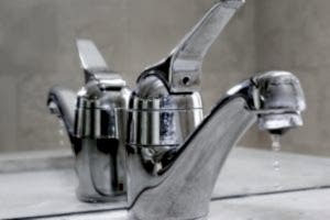 Close-up of a chrome bathroom faucet with tap dripping water, highlighting the issue of a leaky tap. The reflection of the faucet in the mirror emphasizes the water droplets, showcasing the need for repair.