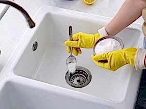Hands in yellow gloves using baking soda to fix a sink that won't drain, showing a DIY unclogging method.