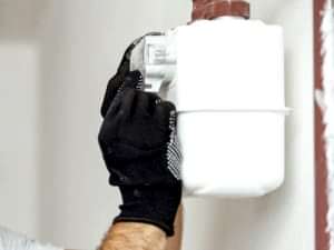 A technician wearing black gloves inspecting a potentially faulty gas meter installed on a white wall.